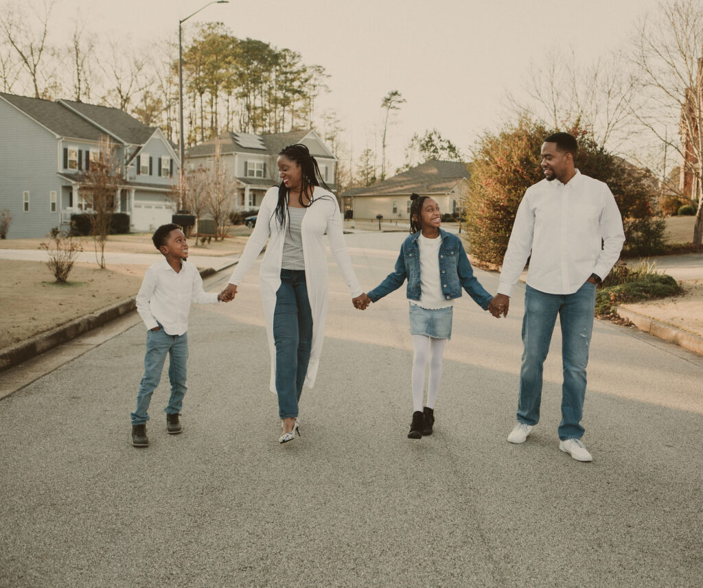 A joyful family of four walks hand-in-hand down a quiet suburban street, smiling at each other. The parents and children wear coordinating outfits with denim and white, enjoying a sunny day together.