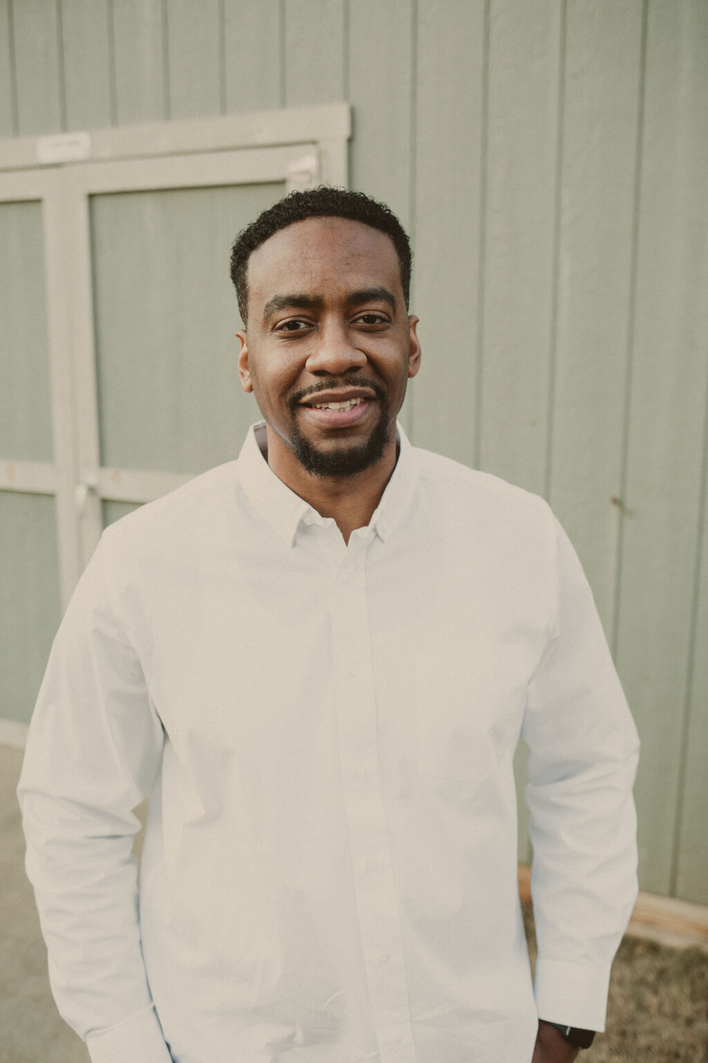 A man in a crisp white button-down shirt stands outdoors in front of a green wooden wall, smiling confidently at the camera.