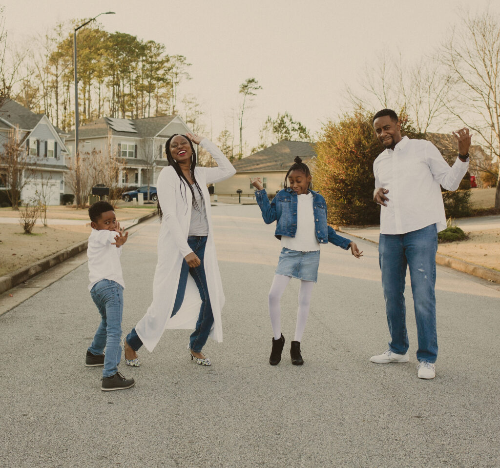 A joyful family of four dances together in the middle of a suburban street, dressed in coordinated casual outfits with white tops and denim. Everyone is smiling and having fun under the afternoon sun.