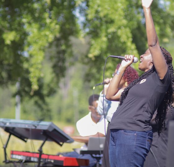 A woman passionately sings into a microphone with one arm raised during an outdoor worship event, surrounded by musicians and trees in the background.